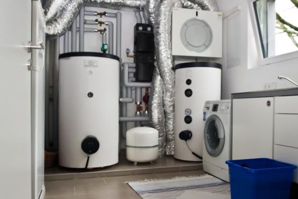 Technikraum mit Wärmepumpe, Pufferspeichern, Lüftungsrohren, Waschmaschine und Trockner auf Fliesenboden.